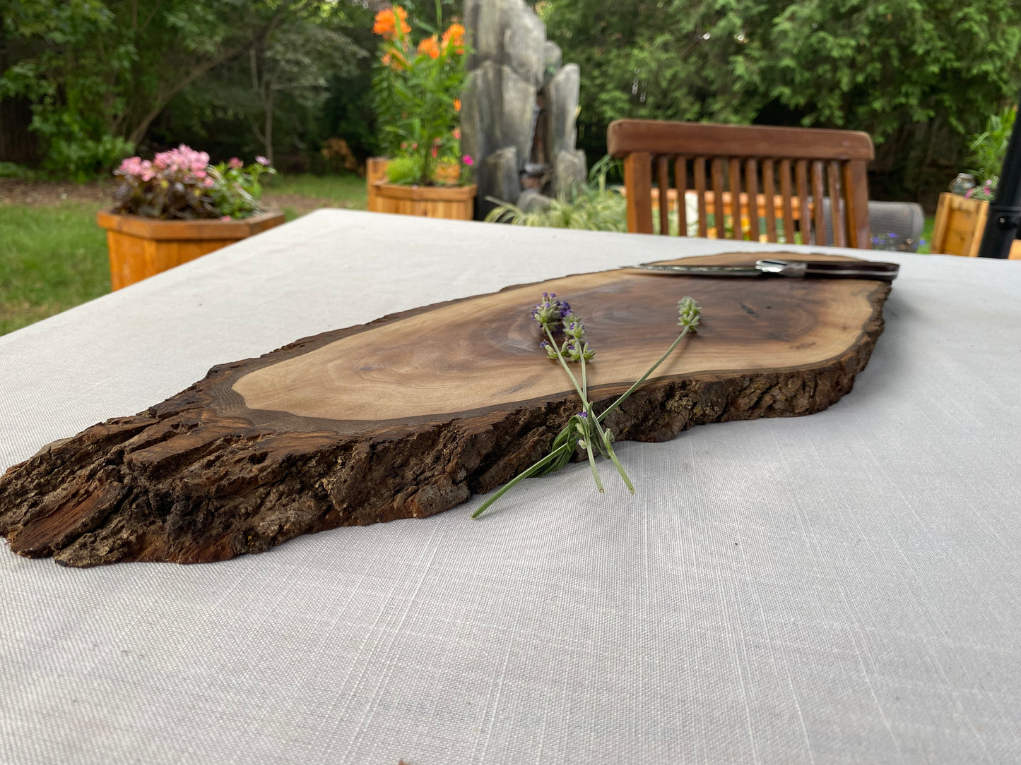 A rustic live-edge wooden serving board with a natural bark border, displayed on a white tablecloth in an outdoor garden setting.