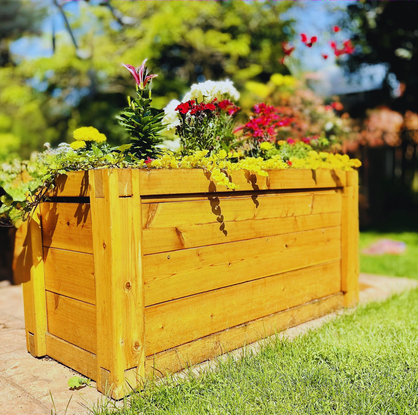 A large rectangular wooden planter box filled with colorful flowers, set outdoors on a brick path in a grassy garden.