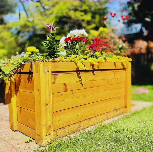 A large rectangular wooden planter box filled with colorful flowers, set outdoors on a brick path in a grassy garden.