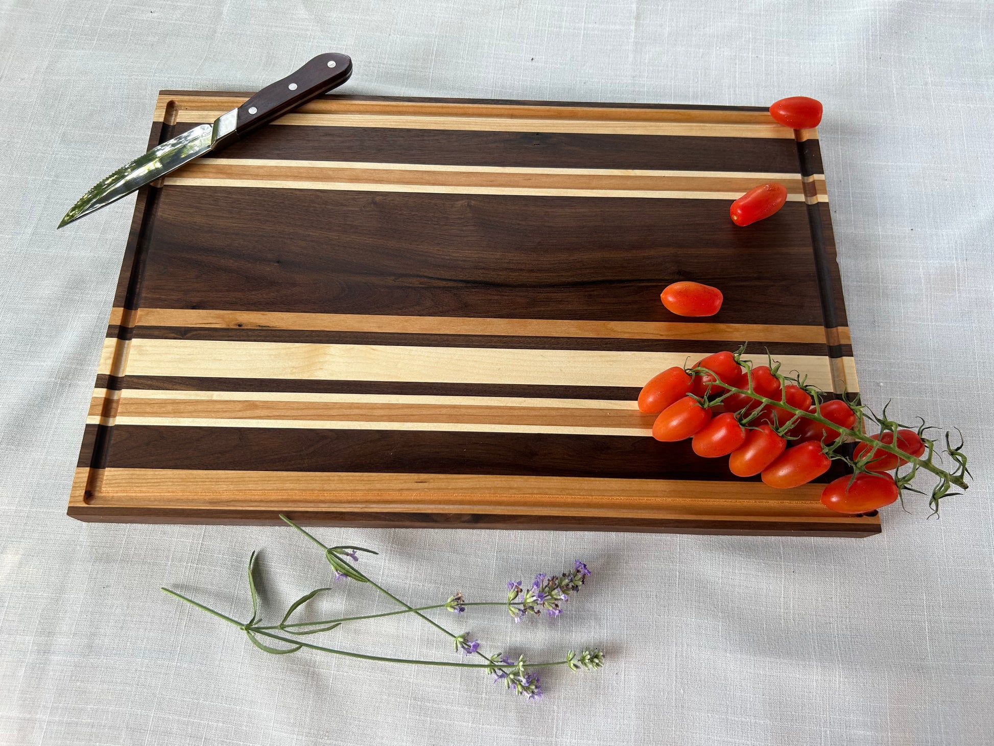 A striped wooden cutting board with alternating light and dark wood bands, styled on a white textured fabric background with a knife, cherry tomatoes, and lavender sprigs.
